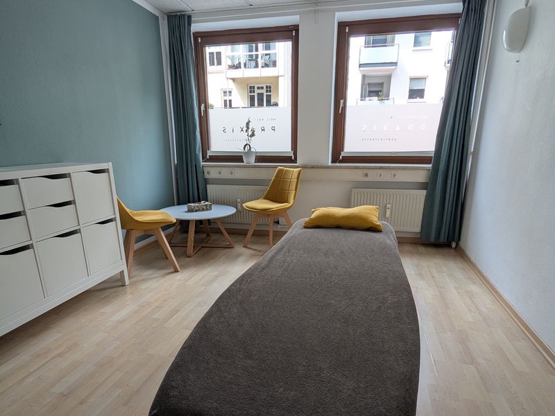 Behandlungsraum mit Liege, zwei gelben Stühlen und Sideboard vor einer petrolfarbenen Wand