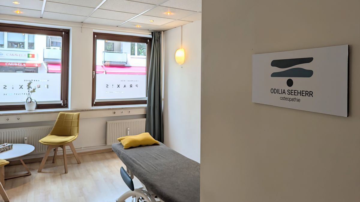 Blick durch die Tür in den Behandlungsraum, links die Behandlungsliege, rechts das Praxisschild Odilia Seeherr Osteopathie